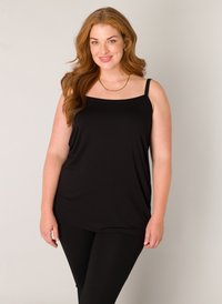 Lachende vrouw met lang rood haar, gekleed in een zwarte mouwloze top, zwarte legging en een gouden ketting, tegen een effen achtergrond.