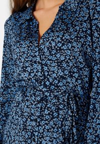 Námorní šaty s květinovým vzorem, které mají zapínání na knoflíky, modro-bílé květinové vzory, dlouhé rukávy a detail s vázáním v pase.