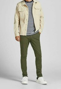 Giacca button-up beige sopra una camicia a righe navy e bianche, abbinata a pantaloni verde olive e sneaker bianchi. Tessuto strutturato, vestibilità casual.