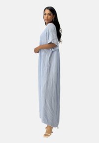 Robe maxi en tissu bleu clair froissé, manches courtes, silhouette fluide et texture subtile. Mannequin debout sur fond neutre.
