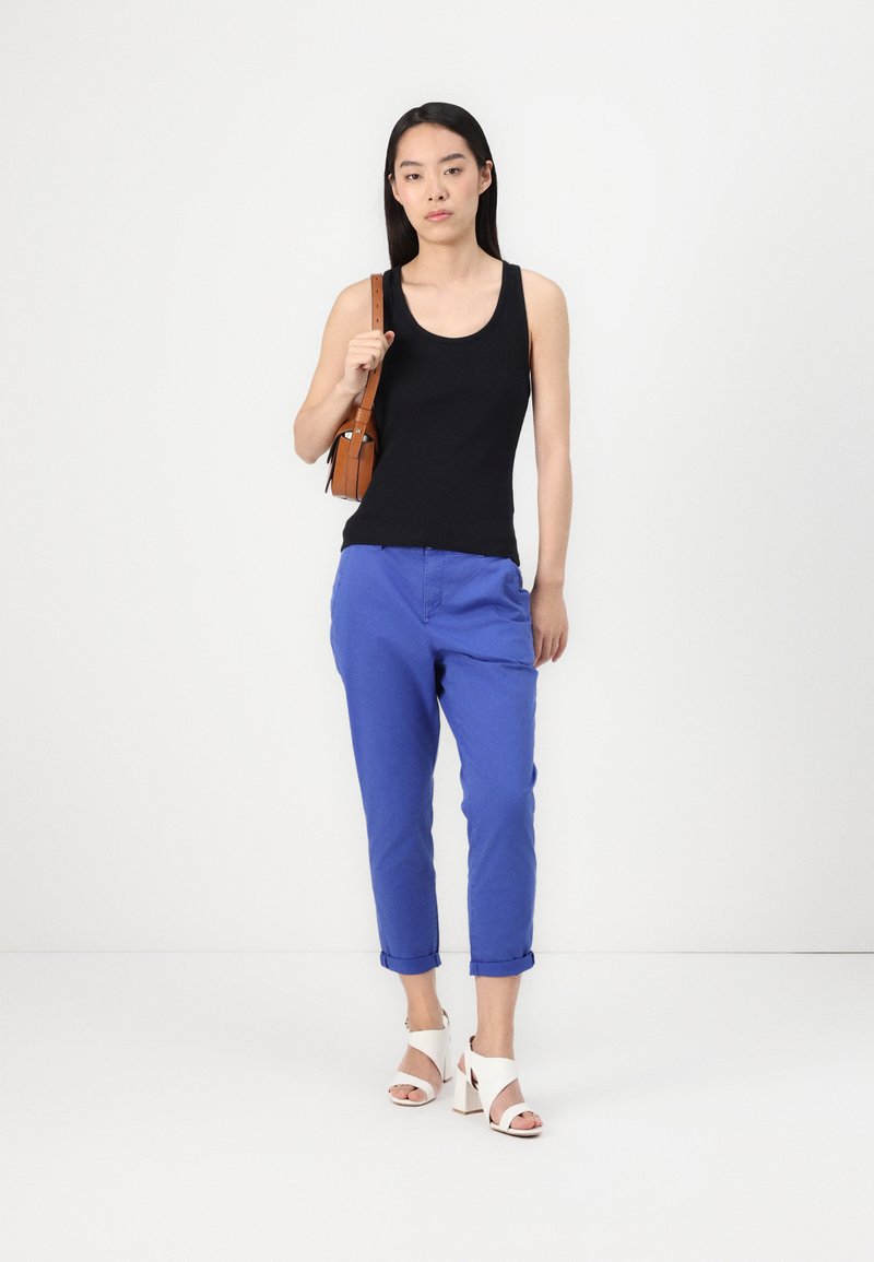 Schwarzes Tanktop, blaue verkürzte Hose, weiße Blockabsatzsandalen und eine braune Umhängetasche, vor einem schlichten weißen Hintergrund positioniert.