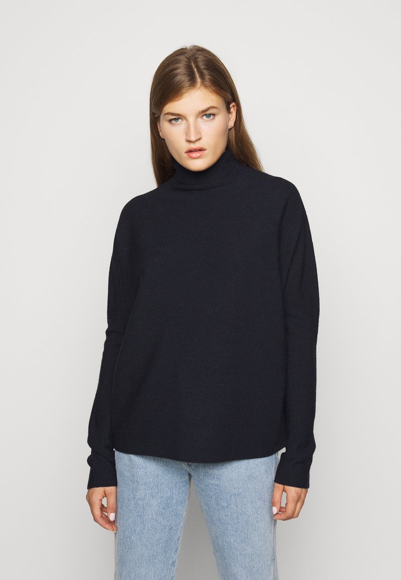 Mørkeblå strømpehalsgenser laget av myk strikket stoff; løs passform med lange ermer og ribbestrikket krage. Kombinert med lyseblå jeans.