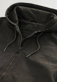 Veste à capuche marron foncé avec fermeture éclair sur le devant, capuche avec cordon de serrage et étiquette visible à l'intérieur du col indiquant la marque et la taille.