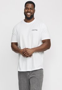 Camiseta de algodón blanca con el texto "Club1990" en negro en el área del pecho izquierdo; combinada con jeans de mezclilla grises.
