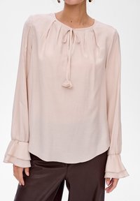 Jasnoróżowa bluzka z długim rękawem, okrągłym dekoltem, zawiązywaniem, plisami i falbaniastymi mankietami, wykonana z miękkiego, lekkiego materiału.