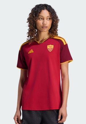 Femme aux cheveux bouclés portant un maillot de sport rouge et jaune avec un logo sur la poitrine, debout devant un fond clair uni.