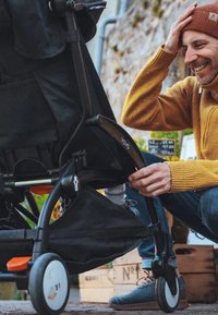 Cochecito negro con un armazón plegable, acentos naranjas y ruedas. Una persona lo ajusta mientras está sentada, con tejidos texturizados visibles.
