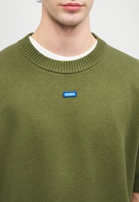 Πράσινο πλεκτό πουλόβερ με ραμμένη λαιμόκοψη και μπλε λογότυπο "HUGO" κεντραρισμένο στο στήθος. Απαλή υφή με χαλαρή εφαρμογή.