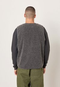 Sivi flis brezrokavnik z teksturirano hrbtno stranjo in temnimi rokavi, kombiniran z zelenimi hlačami. Ima zaobljen izrez in rebraste manšete.