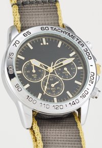 Chronographur med sort urskive, sølv bezel, gule detaljer og grå vævet rem, der indeholder en tachymeter og tre subure.