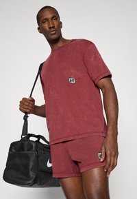 Conjunto de camiseta y pantalones cortos de algodón color burdeos con una etiqueta de logo en el pecho. El modelo lleva una bolsa de deporte negra de Nike con detalles de malla.