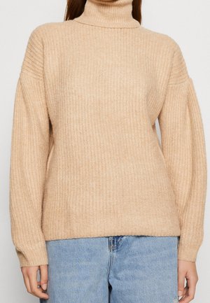 Pull beige à col roulé en tricot côtelé avec manches longues bouffantes, porté avec un jean en denim bleu clair.