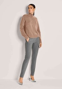 Frau, die einen braunen Strickpullover, graue schmale Hosen mit Seitentasche und silberne hohe Absätze trägt, steht vor einer blassen Wand.