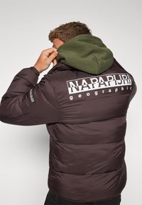 Puffjacka i mörkbrunt med huva i grön, med en stor vit logotyp på baksidan och en texturerad, isolerad design.