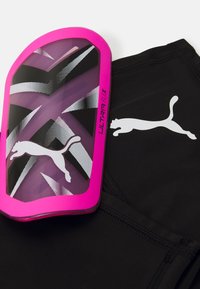 Le protège-tibia Puma présente une coque en plastique rose et violet avec un design géométrique et le texte "ULTRA FLEX", associé à un short en tissu noir.
