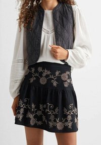 Prošívaná černá vesta přes bílou, strukturovanou blůzu. Černá sukně s jemným zlatým květinovým vyšíváním podél lemu.