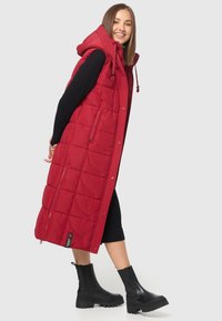 Gilet matelassé rouge avec une capuche, présentant un design en matelassage et des poches latérales. Porté sur une robe noire en côte et associé à des bottes noires.