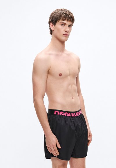 Pantaloni scurți de înot negri, cu o talie elastică lată pe care scrie „DSQUARED2” în roz aprins. Țesătura pare ușoară și fină.