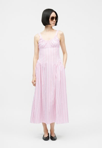 Žena s krátkymi čiernymi vlasmi, ktorá má na sebe veľké slnečné okuliare, ružovo-červené pruhované maxi šaty bez rukávov a čierne topánky, stojaca s jednou rukou v vrecku.