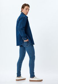 Chaqueta de denim azul oscuro con mangas largas, combinada con jeans ajustados azules. Zapatillas blancas con suelas marrones. Textura suave, ajuste casual.