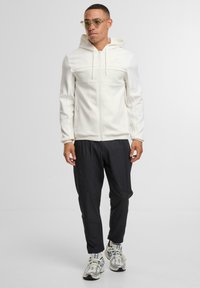 Felpa bianca con zip e pannello in rete, cappuccio con cordino, abbinata a pantaloni jogger neri e scarpe da ginnastica grigie con accenti blu. Indossati occhiali con montatura trasparente.