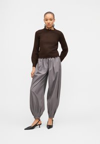 Γυναίκα με πλεγμένα μαλλιά που φοράει σκουρό καφέ πουλόβερ με μακριά μανίκια, χαλαρό γκρι παντελόνι με πιέτες και μαύρα μυτερά γόβες με λουρί στο πίσω μέρος.