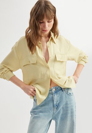 Light yellow button-up shirt with two front pockets, relaxed fit, and long sleeves, paired with light blue high-waisted jeans.