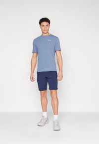Lys blå t-skjorte med korte ermer og en liten hvit logo, kombinert med marineblå shorts. Modellen har på seg grå sneakers med hvite sokker.