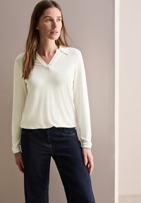 Top bianco a maniche lunghe con scollo a V e tessuto morbido, abbinato a jeans blu scuro con dettagli di cucitura in evidenza.