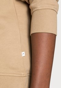 Primo piano di un braccio di carnagione scura che indossa un indumento a maniche lunghe beige con un piccolo etichetta bianca contrassegnata con "/F" vicino alla cucitura.
