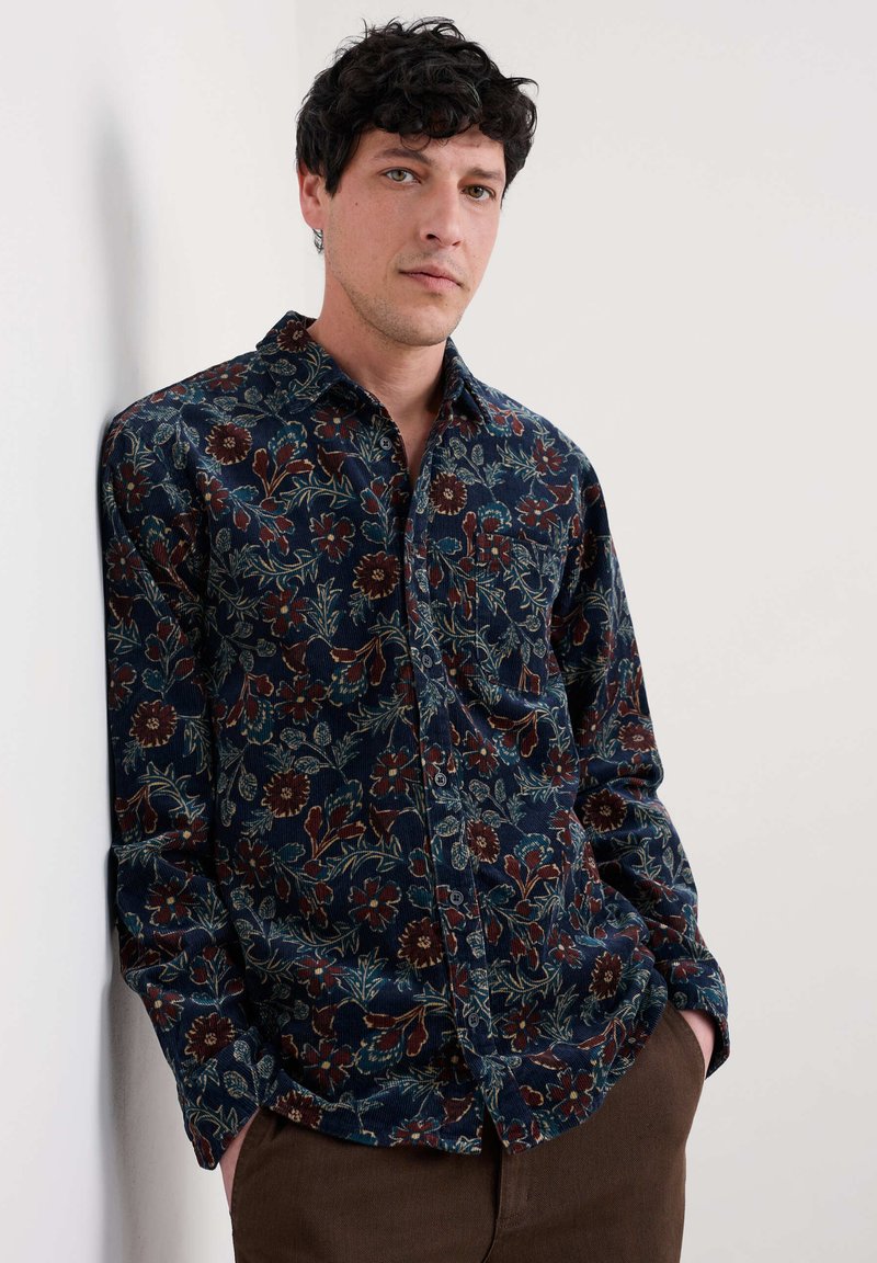 Tums ziedu raksta pogu krekls tumši zilā krāsā ar sarkanbrūniem un zaļiem akcentiem, izgatavots no teksturētas auduma, ar krūšpogu un garām piedurknēm.