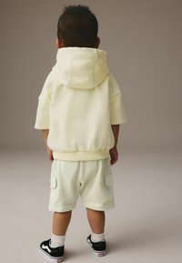 Haut à capuche jaune clair avec une grande capuche, des manches courtes et un bas élastique. Shorts assortis jaunes clairs avec des poches, portés avec des baskets noires.