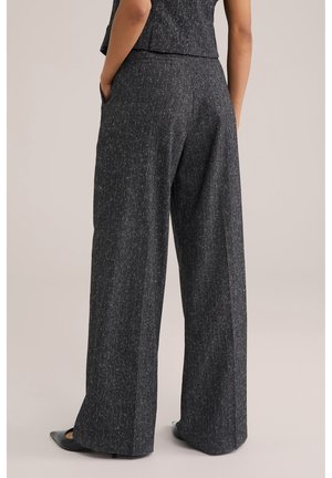 Persoon draagt antracietgrijze wijde broek met subtiele krijtstrepen, handen in de zakken, gecombineerd met zwarte puntige schoenen.