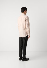 Helles rosa Langarmhemd mit Rückenpasse, geschwungenem Saum und dezentem Muster. Kombiniert mit schwarzen Hosen und Schuhen, von hinten betrachtet.