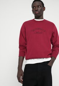 Sweatshirt bordeaux avec col rond, présentant un texte brodé "ORIGINALS". Superposé à une chemise blanche. Matériau en coton doux ; coupe décontractée.