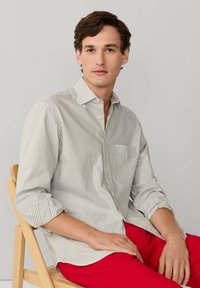 Chemise à rayures boutonnée en vert clair et blanc, tissu texturé, avec une poche poitrine et des manches retroussées. Modèle assis sur une chaise en bois.