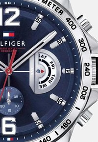 Orologio con quadrante blu, contrassegni argentati, grandi numeri bianchi e una lancetta dei secondi rossa; include la visualizzazione del giorno della settimana e una sottile texture.