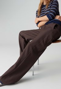 Personne assise sur une chaise portant un pantalon marron à jambes larges et un pull rayé bleu marine, les bras croisés.