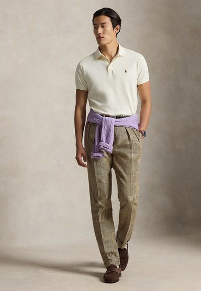 Muž nosící bílé polo tričko, hnědé kárované kalhoty, světle fialový svetr uvázaný kolem pasu, hnědé mokasíny a hodinky, stojící na neutrálním pozadí.