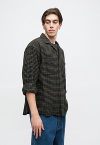 Camisa xadrez preta e marrom com fecho de botões, punhos dobráveis e dois bolsos no peito, usada por cima de uma camisa cinza e combinada com jeans azuis.