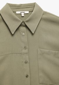 Camicia verde olivo con colletto, sei bottoni e taschino sul petto. Realizzata in tessuto testurizzato con dettagli di cucitura visibili.