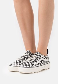 Czarno-białe szachownicowe trampki z płóciennego materiału o teksturowanej powierzchni, kremową gumową podeszwą i kontrastującymi czarnymi akcentami. Sznurowane beżowymi sznurówkami.