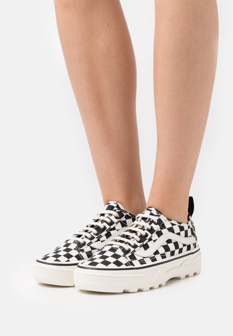 Czarno-białe szachownicowe trampki z płóciennego materiału o teksturowanej powierzchni, kremową gumową podeszwą i kontrastującymi czarnymi akcentami. Sznurowane beżowymi sznurówkami.