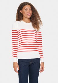 Maglione a righe rosse e bianche con collo rotondo, polsini e orlo a coste. Indossato con jeans blu scuro, caratterizzati da una consistenza morbida.