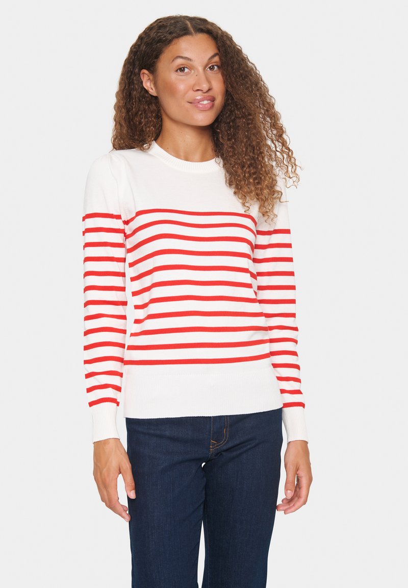 Maglione a righe rosse e bianche con collo rotondo, polsini e orlo a coste. Indossato con jeans blu scuro, caratterizzati da una consistenza morbida.