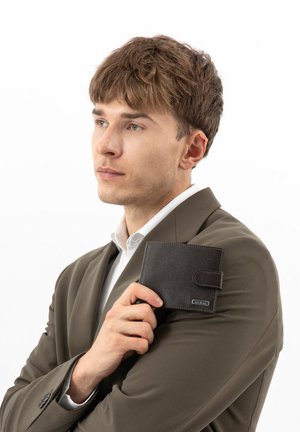 Jonge man in een bruin pak die een zwarte leren portemonnee bij zijn kin houdt en bedachtzaam naar de zijkant kijkt tegen een witte achtergrond.