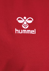 Wit gestileerd bijenlogo met de tekst "hummel" geborduurd op een rode, gestructureerde stofachtergrond.