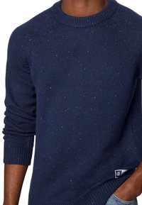 Man med en marinblå melerad stickad tröja med ribbade muddar och rund hals, med handen i fickan, avskuren vid bröstet och höfterna.