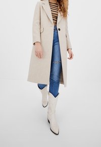 Abrigo beige de lana con cuello con muesca, botón negro único, combinado con vaqueros azules rotos y botas vaqueras blancas con patrones en relieve.