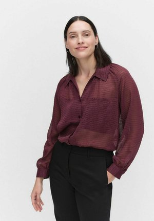 Chemise en Bourgogne, transparente, avec un motif texturé, une poche simple et des manches longues. Associée à un pantalon noir.